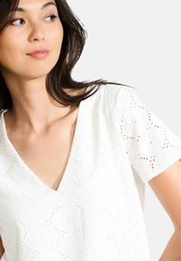 Balta eyelet blūze ar V veida kaklu, īsām piedurknēm un sarežģītām apļa formām. Mīksta tekstūra, viegla materiāla, izceļot detalizētus izgriezumus.