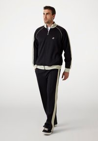 Survêtement noir avec des rayures blanches sur les côtés, comprenant une veste zippée et un pantalon assorti. Fabriqué en tissu lisse avec un design sportif.