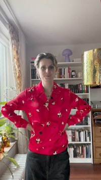 Czerwona bluzka w kwiatowy wzór, z długimi rękawami i kołnierzykiem, zestawiona z ciemnymi dżinsami. W tle regał z książkami i dekoracyjna lampa.
