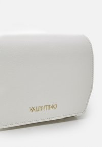 Vit läderhandväska med en slät textur, guldlogo "VALENTINO" och en strukturerad form. Har rena linjer och en minimalistisk design.