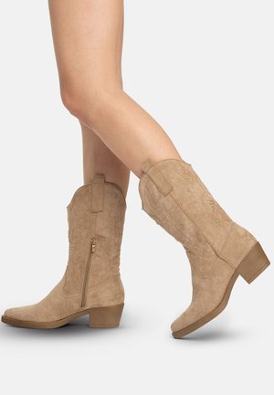 Benedenbenen met beige suède westernlaarzen tot halverwege de kuit, met lage blokhakken en zijritsen, op een witte achtergrond.