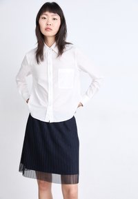 Camisa blanca a rayas con botones y un bolsillo en el pecho, combinada con una falda plisada en color marino que presenta un detalle de dobladillo de malla.