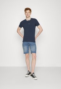 Tamsiai mėlynas trumpomis rankovėmis polo marškinėliai su baltais akcentais, derinami su šviesiai mėlynomis džinsinėmis šortais ir juodai baltais sportbačiais.