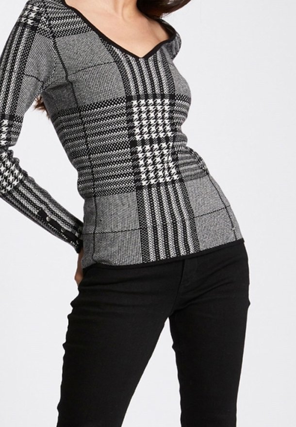Femme portant un haut ajusté à manches longues à carreaux noirs et gris avec un col en V, assorti à un pantalon noir, debout devant un fond uni.
