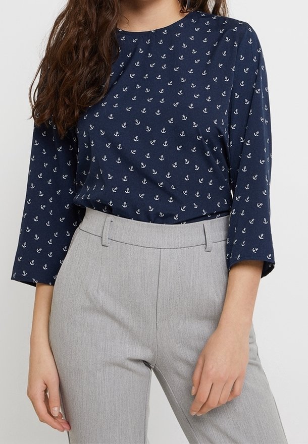 Femme portant un haut bleu marine à manches longues avec des motifs d'ancres blanches rentré dans un pantalon taille haute gris clair.