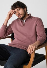 Világos rózsaszín, cipzáros pulóver, bordázott mandzsettával és aljjal, fehér ing fölött viselve, sötét farmernadrággal párosítva. A modell egy fából készült széken ül.