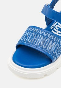 Blå sandaler med en texturerad tygrem med ett vitt logomönster, vadderad sula och en kraftig vit gummisula.