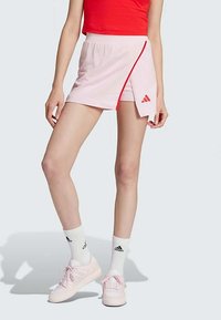 Saia desportiva rosa com uma fenda lateral, apresentando um detalhe em vermelho e um logo bordado da Adidas. Usada com meias brancas e ténis rosa.