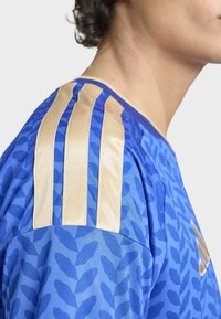 Blå träningsskjorta med ett strukturerat lövmönster, gyllene tre-randiga detaljer på axlarna och rund halsringning.