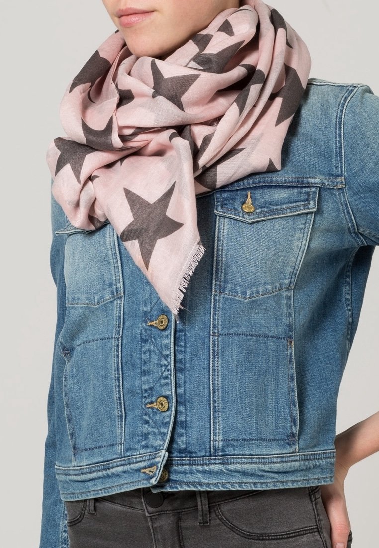 Personne portant une écharpe rose clair avec de grandes étoiles foncées enroulée autour du cou et une veste en jean bleue avec des boutons en laiton.