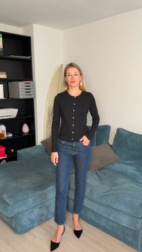 Γυναίκα με ξανθά μαλλιά που φοράει μαύρη ζακέτα με κουμπιά, μπλε τζιν και μαύρες γόβες, στέκεται σε ένα σαλόνι με μπλε καναπέ και ράφια.