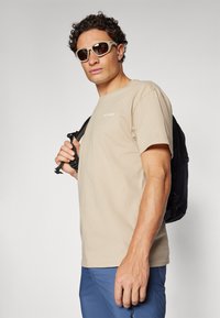 Tricou bej cu mânecă scurtă și logo alb, asortat cu pantaloni albaștri. Persoana poartă ochelari de soare maro și are în spate un rucsac negru.