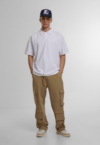 Hvit polo skjorte, beige cargo bukser med sidelommer, og beige joggesko. Tilbehør: blå caps med hvit logo, klare briller. Nøytral bakgrunn.