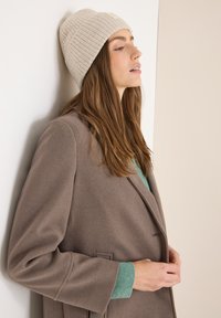 Abrigo de lana marrón sobre un suéter verde azulado, combinado con un gorro de punto beige acanalado que tiene una pequeña etiqueta. El cabello es largo y liso.