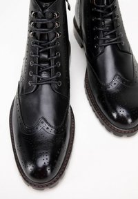 Černé kožené kotníkové boty s wingtip designem, dekorativními perforacemi a kulatými tkaničkami. Silná gumová podrážka s texturovaným povrchem.