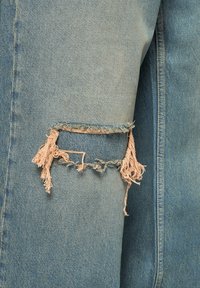 Veste en denim bleu clair avec une manche usée. Bords effilochés avec des fils lâches au niveau du coude, soulignant une texture usée.