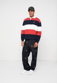Man staat in een gestreepte rugbyshirt in rood, wit en marineblauw, donkere jeans met geborduurde logo's, witte sneakers en een zwarte pet.