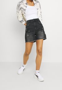 Shorts de mezclilla negra con dobladillo deshilachado, detalles desgastados, diseño de talle alto, combinados con un top blanco sin mangas y una camisa teñida con efecto tie-dye. Zapatillas blancas.