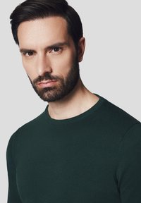 Ciemnozielony sweter dzianinowy z okrągłym dekoltem i dopasowanym krojem. Gładka tekstura z delikatnym rozciągnięciem. Zbliżenie na górną część ciała i twarz.