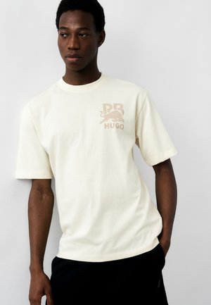 Νεαρός άνδρας που φοράει μια χαλαρή κρεμ μπλούζα με λογότυπο ταν λέοντα και κείμενο "PB HUGO" στο στήθος, στέκεται μπροστά σε απλό φωτεινό φόντο.