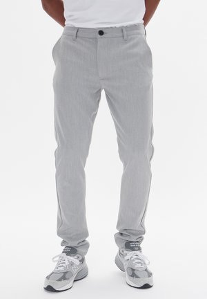 Homme portant un chino gris clair et des baskets New Balance grises, debout, les mains dans les poches, devant un fond blanc.