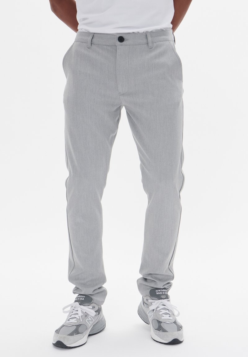 Homme portant un chino gris clair et des baskets New Balance grises, debout, les mains dans les poches, devant un fond blanc.