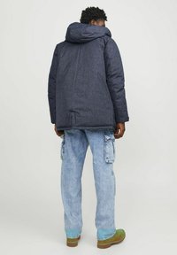Chaqueta acolchada azul oscuro con una gran capucha, que presenta un tejido texturizado. Llevada sobre pantalones cargo azul claro y zapatos verdes.