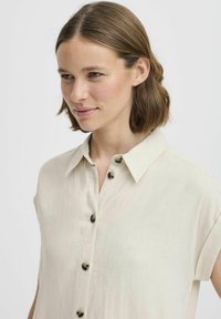 Camicia beige con colletto a maniche corte, dotata di una chiusura a bottoni sul davanti ornata da bottoni scuri e realizzata in un tessuto leggero e testurizzato.