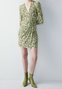 Robe fleurie en vert et marron, avec des manches longues, un décolleté en V profond, une taille froncée et une longueur courte, associée à des bottines vertes.