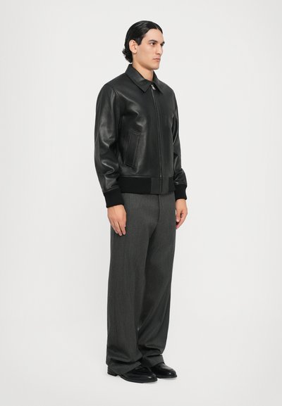 Veste bomber en cuir noir avec une fermeture éclair à l'avant, un col et des poignets côtelés, associée à un pantalon large gris et des chaussures noires.