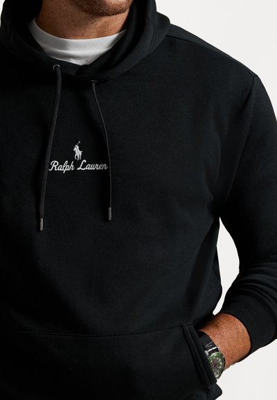 Mężczyzna w czarnej bluzie z kapturem Ralph Lauren z białym logo, białej koszulce oraz zielonym zegarku na lewej nadgarstku.