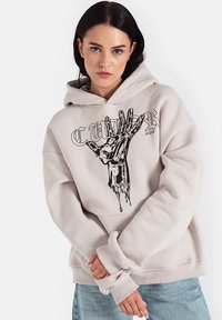 Beżowy hoodie z dużą czarną grafiką dłoni z przodu, charakteryzujący się luźnym krojem, kapturem ze sznurkiem i ściągaczami na rękawach. Pod spodem znajduje się jasnoniebieska koszulka.