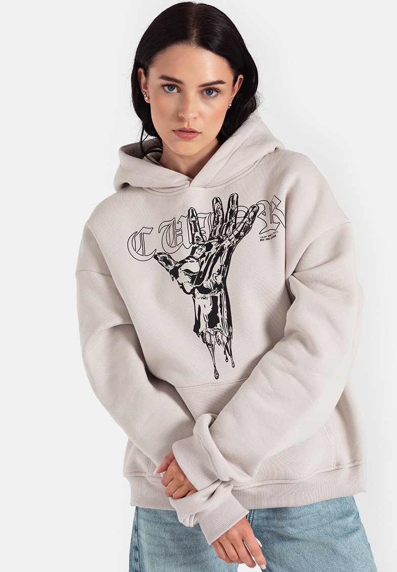 Beżowy hoodie z dużą czarną grafiką dłoni z przodu, charakteryzujący się luźnym krojem, kapturem ze sznurkiem i ściągaczami na rękawach. Pod spodem znajduje się jasnoniebieska koszulka.