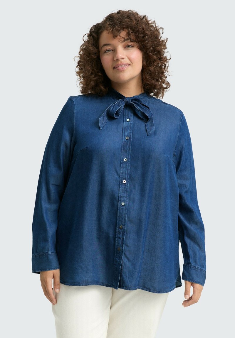 Niebieska dżinsowa koszula z długimi rękawami, z dopasowanym kołnierzykiem i muszką przy szyi. Posiada zapięcie na guziki z przodu oraz miękką teksturę materiału.