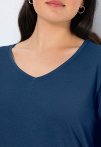 Kobieta nosząca srebrne kolczyki w kształcie kół oraz niebieską koszulę z dekoltem w serek, eksponująca swoją szyję i dolną część twarzy na jasnym tle.