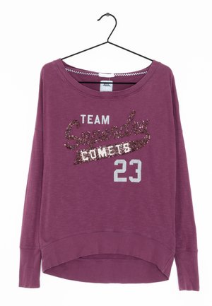 Lila Sweatshirt mit langen Ärmeln an einem schwarzen Kleiderbügel, mit dem Schriftzug "TEAM Superdry COMETS 23" aus weißen und braunen Pailletten auf der Vorderseite.
