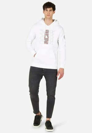 Felpa bianca con stampa grafica e cordoncino, indossata con jeans neri aderenti e sneaker nere con suola bianca. Design minimalista, abbigliamento casual.