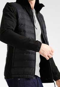 Man som bär en svart huvaquiltad jacka över en grå henley-tröja och mörkblå jeans, som justerar dragkedjan på jackan.