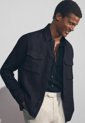 Camicia nera a maniche lunghe in morbido tessuto, con due taschini sul petto, colletto classico e taglio rilassato, abbinata a pantaloni chiari.