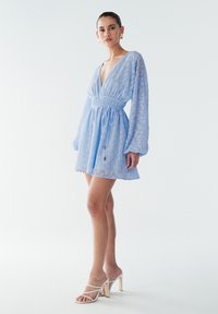 Svetlo modra obleka z dolgimi, voluminoznimi rokavi, globokim v-izrezom in nabranim pasom. Prosojen material ima subtilen vzorec. Stilizirano z belimi petami.