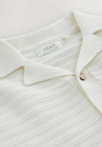 Vit stickad pikétröja med krage, en enda knapp i trä och strukturerade horisontella ränder. Etiketten visar "NEXT, tillverkad i Bangladesh."