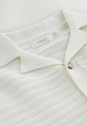 Weißes Strick-Poloshirt mit einem Kragen, einem einzelnen Holzknopf und strukturierten horizontalen Streifen. Das Etikett trägt die Aufschrift "NEXT, hergestellt in Bangladesch."