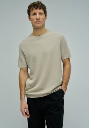 Hombre de pie con las manos en los bolsillos, vestido con una camiseta de punto beige de manga corta y pantalones negros sobre un fondo liso.