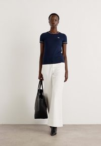 T-shirt ajusté bleu marine avec un col contrastant rouge et blanc, associé à un pantalon blanc évasé et à un sac fourre-tout noir, sur un fond neutre.