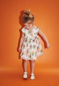 Weißes Kleid mit buntem Blumenstickerei, kurzen Ärmeln, tailliertem Bund und ausgestellt Rock, kombiniert mit weißen Schuhen. Hintergrund ist orange.