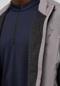 Leichte graue Jacke mit einem weichen, kuscheligen schwarzen Innenfutter; getragen über einem dunkelblauen Oberteil mit einem halben Reißverschlusskragen. Strukturierten Stoffdetails sichtbar.