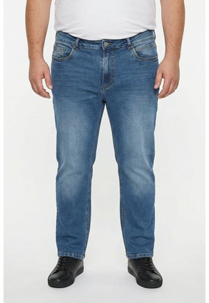 Mann trägt mittelblaue Jeans, weißes T-Shirt und schwarze Schnürsneaker, steht vor einem einfarbigen hellen Hintergrund.