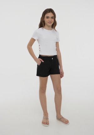 T-shirt bianco corto con ritagli laterali, abbinato a pantaloni neri con vita elasticizzata e tasca. Sandali grigi completano il look.