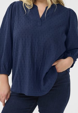 Femme portant un chemisier texturé bleu marine avec manches 3/4 et un jean bleu foncé, main dans la poche, torse visible.