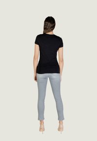 Femme aux longs cheveux bruns portant un haut noir ajusté à manches courtes, un jean skinny gris clair et des chaussures à talons hauts beiges, debout de dos.